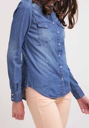 Femme portant une chemise en jean bleue cintrée à boutons et un pantalon beige clair, photographiée du cou jusqu'aux hanches, les bras détendus le long du corps.