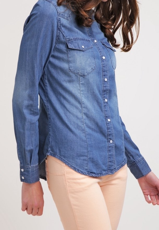 Femme portant une chemise en jean bleue cintrée à boutons et un pantalon beige clair, photographiée du cou jusqu'aux hanches, les bras détendus le long du corps.