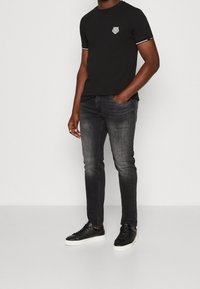Camiseta negra de manga corta con emblema en el pecho, combinada con jeans oscuros de corte slim y zapatillas negras con suelas blancas.
