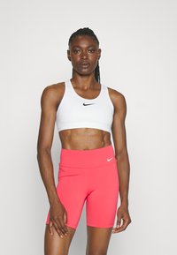 Biały stanik sportowy z czarnym logo Nike w połączeniu z różowymi szortami o wysokim stanie. Gładki, elastyczny materiał z dopasowanym krojem.