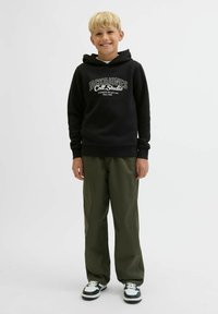 Smaidošs blond zēns stāv melnā "Jack & Jones" kapucē, olīvzaļa biksēm un melni-baltām sporta kurpēm uz viendabīgas fona.