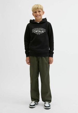Glunderende blonde jongen die staat in een zwarte 'Jack & Jones' hoodie, olijfgroene broek en zwart-witte sneakers voor een effen achtergrond.
