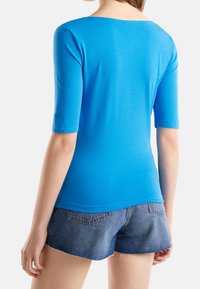 Modrá košile s krátkými rukávy a kulatým výstřihem, hladká textura, přiléhavý střih a jemné švy, v kombinaci s džínovými šortkami.