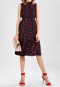 Robe noire avec imprimés en forme de cœur rouges, design sans manches, taille cintrée avec une ceinture, ourlet volanté. Assortie à des talons rouges à lanières et à une pochette rose clair.