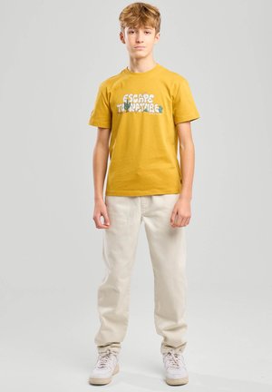 Jugendlicher Junge steht, trägt ein gelbes T-Shirt mit Grafik und Text, hellbeige Hosen und weiße Turnschuhe vor einem schlichten Hintergrund.