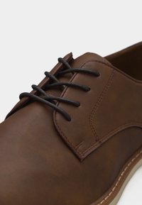 Chaussure habillée en cuir marron avec des lacets marron foncé et des coutures détaillées sur fond blanc, gros plan sur l'avant et le côté.