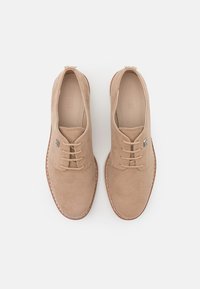 Chaussures habillées en daim beige avec un design à lacets, bout rond, semelle marron contrastante, et un détail discret de logo sur le côté gauche.