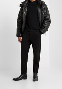 Homme portant une veste matelassée en cuir noire, un pull noir, un pantalon noir fuselé et des bottines en cuir noir texturé, debout devant un fond blanc uni.