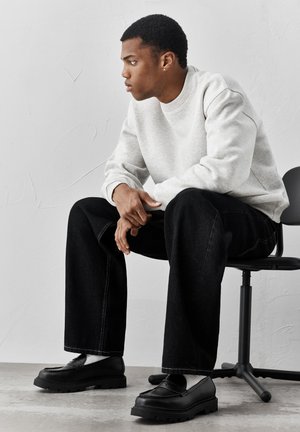 Mladý muž v světle šedé mikině, černých kalhotách s širokými nohavicemi a černých mokasínách sedí na kancelářské židli a dívá se stranou.