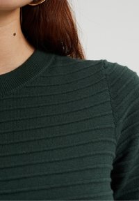 Mörkgrön ribbad stickad tröja med rundad hals och texturerade detaljer, som framhäver en figurnära silhuett och mjukt material.