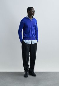 Άνδρας που φορά μπλε πουλόβερ πάνω από λευκό πουκάμισο με γιακά, μαύρο παντελόνι, μαύρα παπούτσια και γυαλιά ηλίου, στέκεται μπροστά σε απλό φωτεινό φόντο.