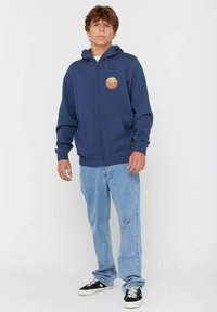 Jonge man die staat in een marineblauwe zip-up hoodie met een cirkelvormige oranje graphic, lichtblauwe spijkerbroek en zwarte sneakers.