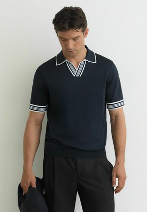 Muž v námornícky modrej košeli s krátkym rukávom, s bielym pruhovaným golierom a manžetami rukávov, drží zodpovedajúcu námornícky modrú čiapku a pozerá sa dole.