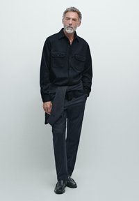 Sort skjorte med knaplukning og to brystlommer, mørke bukser og sorte sko. En grå sweater er bundet om livet. Minimalistisk design.