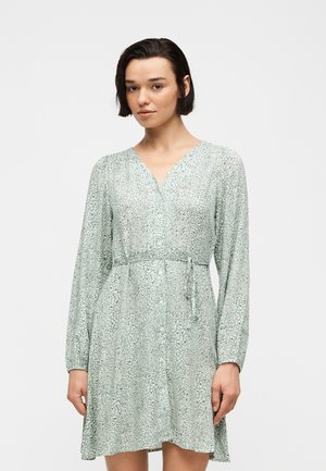 Γυναίκα με κοντά σκουρόχρωμα μαλλιά, που φοράει ένα μακρυμάνικο, ανοιχτοπράσινο φόρεμα με σχέδια, V-λαιμόκοψη και ζώνη στη μέση, στέκεται μπροστά σε απλό λευκό φόντο.