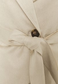 Beżowa kurtka z teksturowanej tkaniny, z opaską przy talii oraz okrągłym brązowym guzikiem z wyraźnymi oznaczeniami.