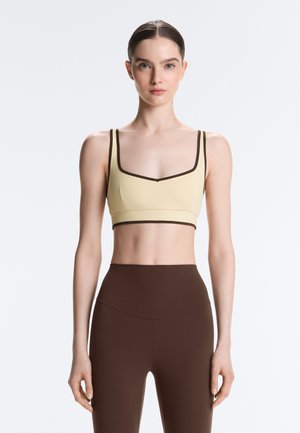 Woman with slicked-back hair wearing a beige crop top with dark trim and high-waisted dark brown leggings, standing against a white background.