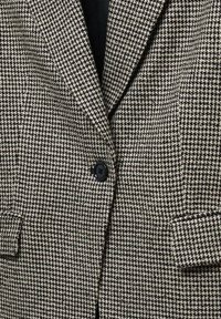 Blazer gris et noir à motif pied-de-poule avec un bouton noir et des poches à rabat, porté sur un top noir.