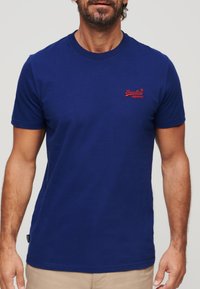 Camiseta de algodón azul marino con mangas cortas, escote redondo y logotipo bordado en rojo en la parte izquierda del pecho. Textura suave, ajuste clásico.