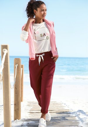 Helles rosa Zip-Hoodie über einem weißen T-Shirt mit farbigem Text. Weinrote Jogginghosen mit Kordelzug und umgeschlagenen Säumen. Weiße Sneakers. Strandumgebung.