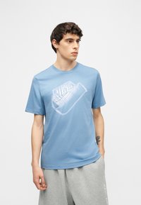 Jeune homme aux cheveux courts et bouclés portant un t-shirt Nike bleu clair et un pantalon de survêtement gris clair, debout devant un fond uni.