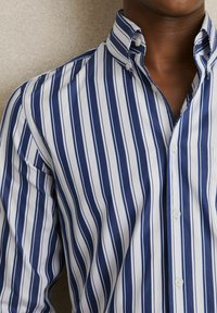 Chemise boutonnée rayée bleu marine et blanche avec un col classique. Présente des rayures verticales, une texture en coton lisse et une poche poitrine unique.