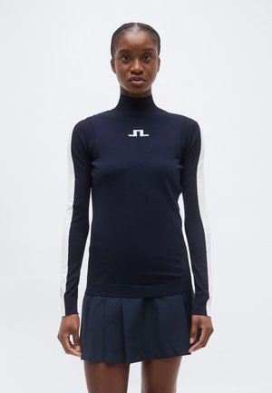 Femme portant un pull à col roulé bleu marine avec des manches blanches et un petit logo blanc, assorti à une jupe plissée bleu marine.