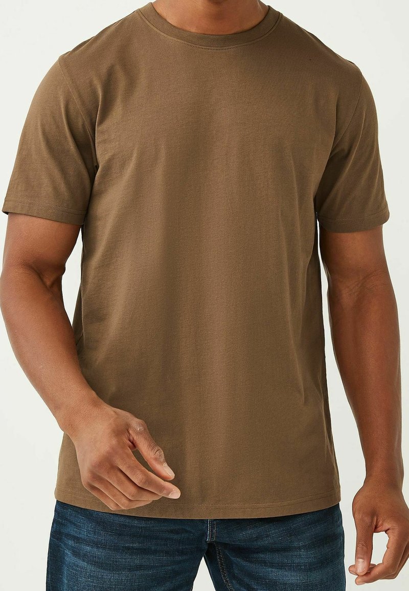 Homme portant un t-shirt marron uni à manches courtes et un jean bleu, vu du cou aux hanches avec une main légèrement levée.