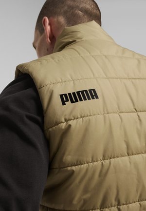 Gilet imbottito beige con cuciture visibili, colletto alto e logo nero sul retro. Indossato sopra una felpa scura.