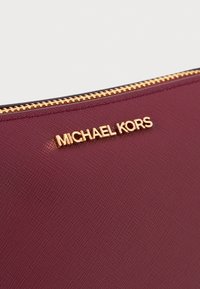 Burgundy- värinen teksturoitu kangaspussi, jossa on kultasävyinen "MICHAEL KORS" -logo ja kultainen vetoketju yläreunassa.