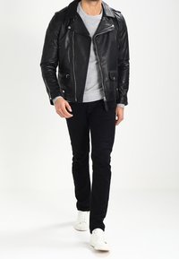 Chaqueta de motociclista de cuero negro con cremalleras plateadas y cuello con muesca, llevada sobre un suéter gris y combinada con pantalones negros y zapatillas blancas.