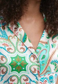 Blusa de satén en blanco con un vibrante patrón paisley en verde, azul y naranja. Presenta un cuello clásico y detalles de botones.