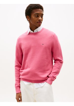 Rosa Strickpullover mit strukturierter Oberfläche, Rundhalsausschnitt und gerippten Bündchen. Ein kleines Logo ist auf der Brust sichtbar, getragen über einem weißen Kragenhemd.