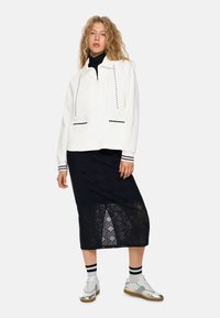 Weißer Pullover mit Reißverschlusskragen und Kordelzügen, kombiniert mit einem strukturierten schwarzen Spitzen-Midi-Rock. Silberne Sneakers und sportliche gestreifte Socken runden den Look ab.