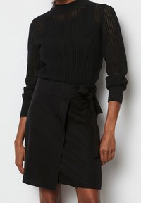 Sort strikket sweater med lange ærmer og høj hals, kombineret med en sort omslagsnederdel med et bindedetalje ved taljen.