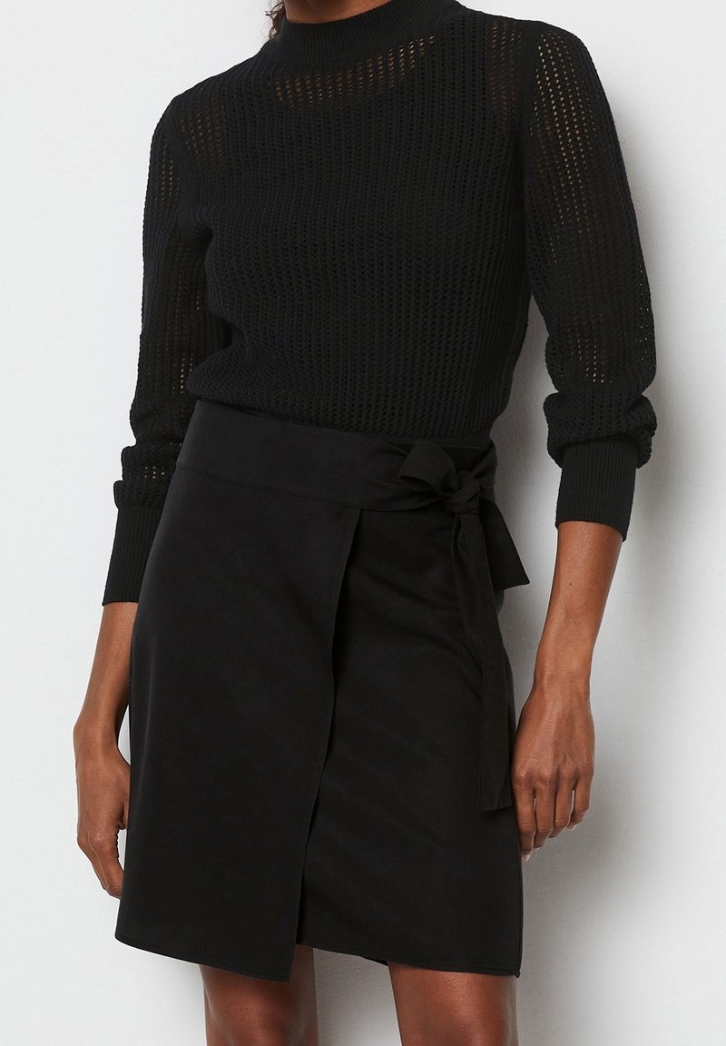 Sort strikket sweater med lange ærmer og høj hals, kombineret med en sort omslagsnederdel med et bindedetalje ved taljen.