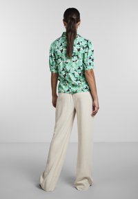 Chemise florale verte à manches courtes, associée à un pantalon large beige. La chemise présente un motif de feuilles vif et une coupe décontractée.