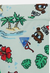Gaiši zils audums ar tropiskām motīvām, kurās redzami palmas, ziedi un multfilmu varoņi. Iekļauts zils trīsstūris ar "GUESS BEACH" logo.