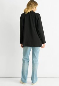 Long black jacket with a straight cut, featuring a smooth fabric and minimal detailing, paired with light blue jeans and beige shoes.