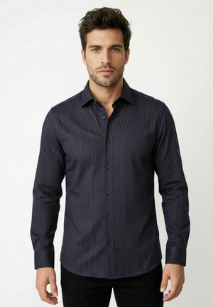 Mann trägt ein enganliegendes, langärmliges, marineblaues Hemd mit dezentem kleinen Punktmuster und steht vor einem schlichten hellen Hintergrund.