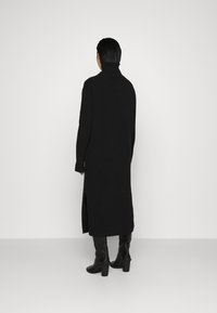 Langes schwarzes Strickkleid mit hohem Kragen, seitlichen Schlitzen und eng anliegenden Ärmeln. Das Modell trägt knielange schwarze Stiefeletten mit Absatz.