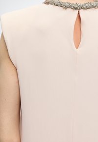 Οπίσθια όψη φόρεμα ρόζ παστέλ χωρίς μανίκια με άνοιγμα σε σχήμα κλειδαρότρυπας και διακοσμητικό κολάρο με στρας στον άνω μέρος της πλάτης και τον ώμο ενός ατόμου.