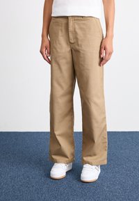 Pantaloni beige a gamba larga in twill di cotone, con frontale piatto e tasche laterali, abbinati a sneakers bianche su un tappeto blu.