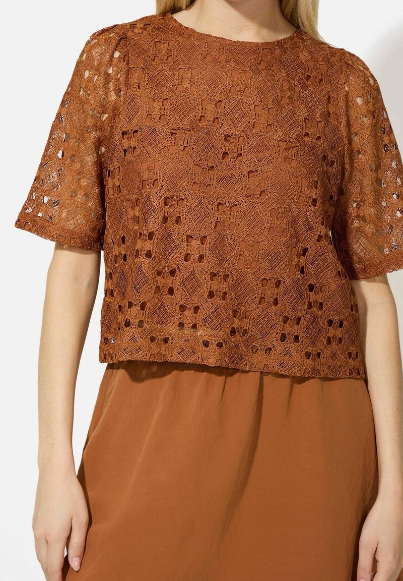 Femme portant un haut en crochet à manches courtes de couleur rouille sur une jupe ou un pantalon assorti de couleur rouille.