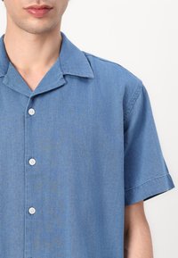 Μπλε τζιν πουκάμισο κοντομάνικο με αιχμηρό γιακά, διαθέτει κουμπιά μπροστά και λείο υλικό. Ορατά είναι τα αντίστοιχα πλαστικά κουμπιά.