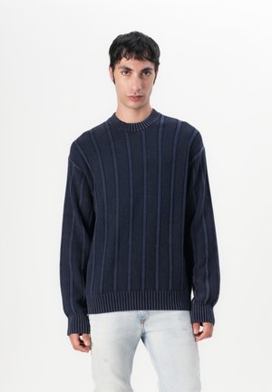 Navy blue knitted sweater with vertical ribbed texture, round neckline, and long sleeves. Paired with light blue distressed jeans.
