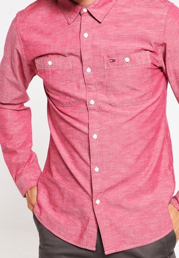 Homme portant une chemise rose à manches longues boutonnée avec deux poches poitrine et un pantalon foncé, les mains dans les poches.