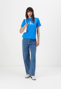 Tricou din bumbac albastru cu logo alb, asortat cu pantaloni din denim bleu deschis cu detalii distresate. Modelul ține o geantă albă mică.