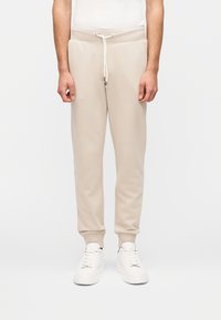 Μπεζ φόρμες με ελαστική μέση με κορδόνι, ριμπ μανσέτες και πλαϊνές τσέπες, κατασκευασμένες από μαλακό, υφασμάτινο ύφασμα με υφή. Φορεμένες με λευκά αθλητικά παπούτσια.