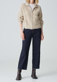 Beige fuzzy zip-up jacket with a front pocket, paired with dark blue wide-leg jeans and brown ankle boots. Simple design, relaxed fit.